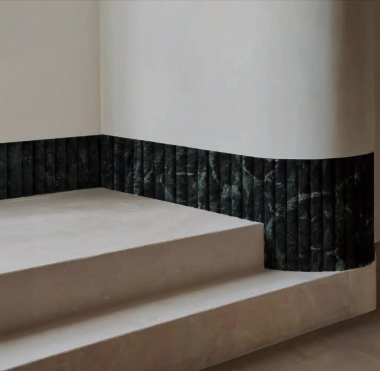 Angled view of minimalist interior stairs with beige concrete steps and a dark, textured marble wainscoting along the lower part of the wall.