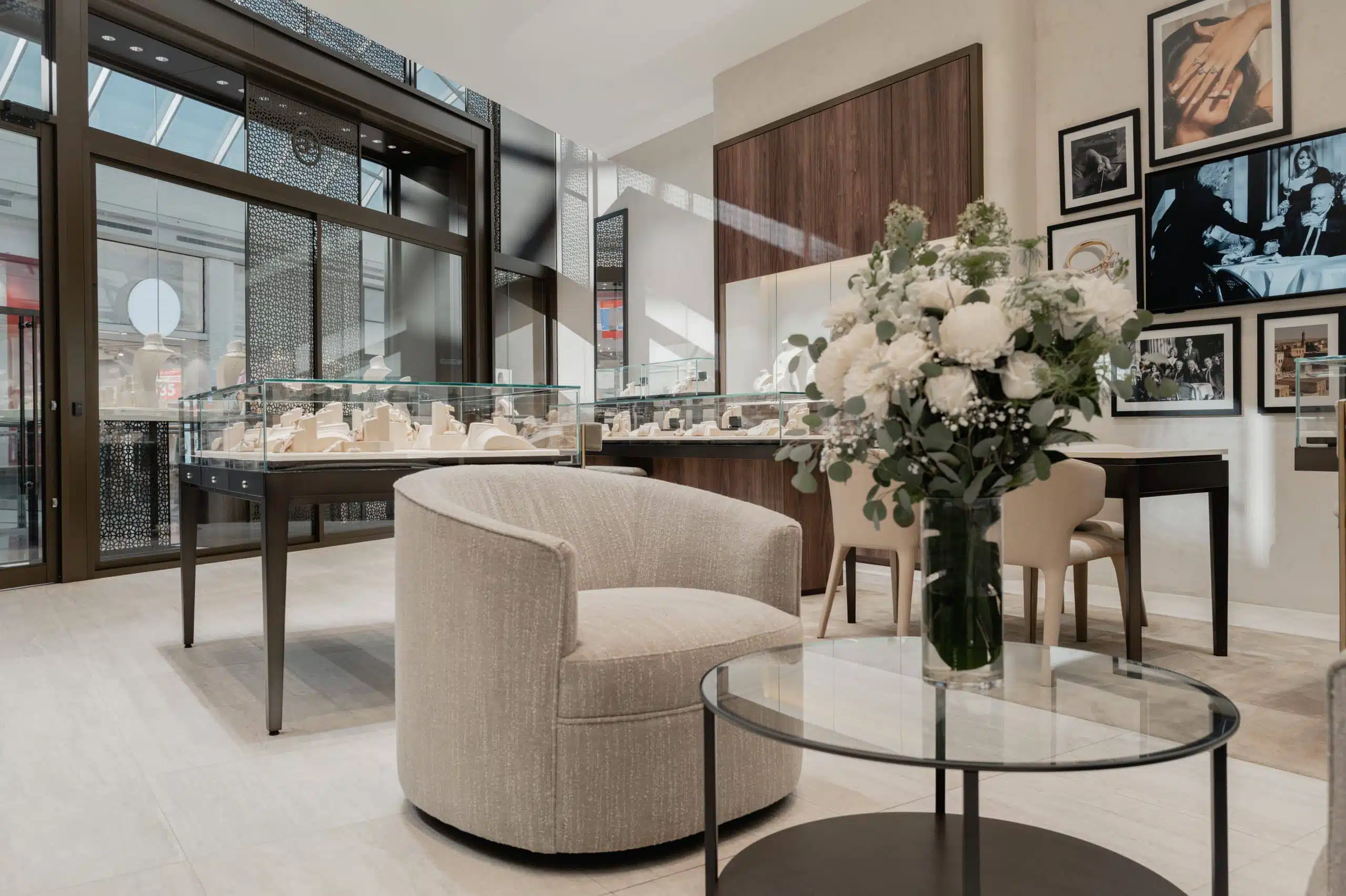 A modern jewelry store interior with glass display cases, a round armchair, a glass table with a bouquet of white flowers, and framed photos on the wall.